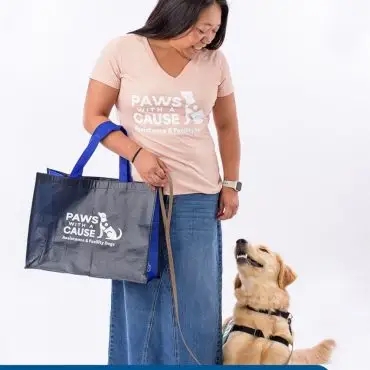 Person with Paws With a Cause shirt holding a bag beside a happy golden retriever, highlighting assistance dogs.