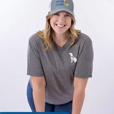 Smiling woman in a Paws for a Cause cap and grey shirt, leaning on a stool.