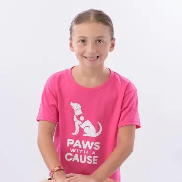 Young girl in a pink Paws with a Cause t-shirt smiles while leaning on a wooden stool.