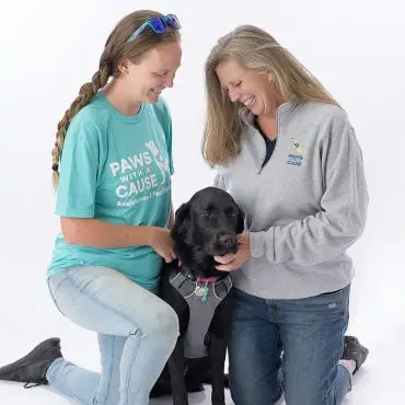 Two smiling women with a black dog in Paws with a Cause shirts, promoting assistance dog programs.