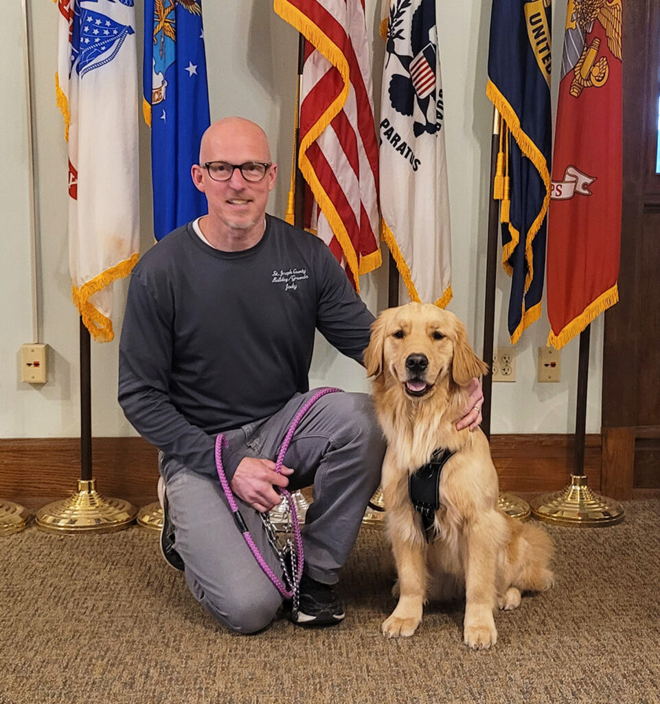 Man kneeling beside happy golden retriever in front of colorful flags indoors.