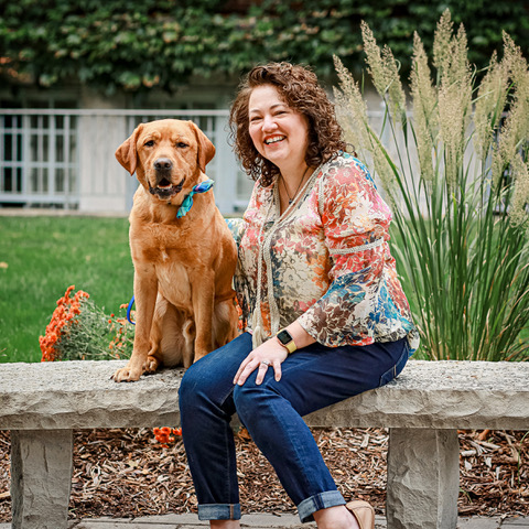 Woman and dog sitting on a stone bench in a garden, both smiling at the camera.