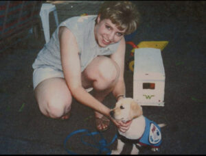 Woman smiling and hugging a Labrador puppy dressed in a blue vest.