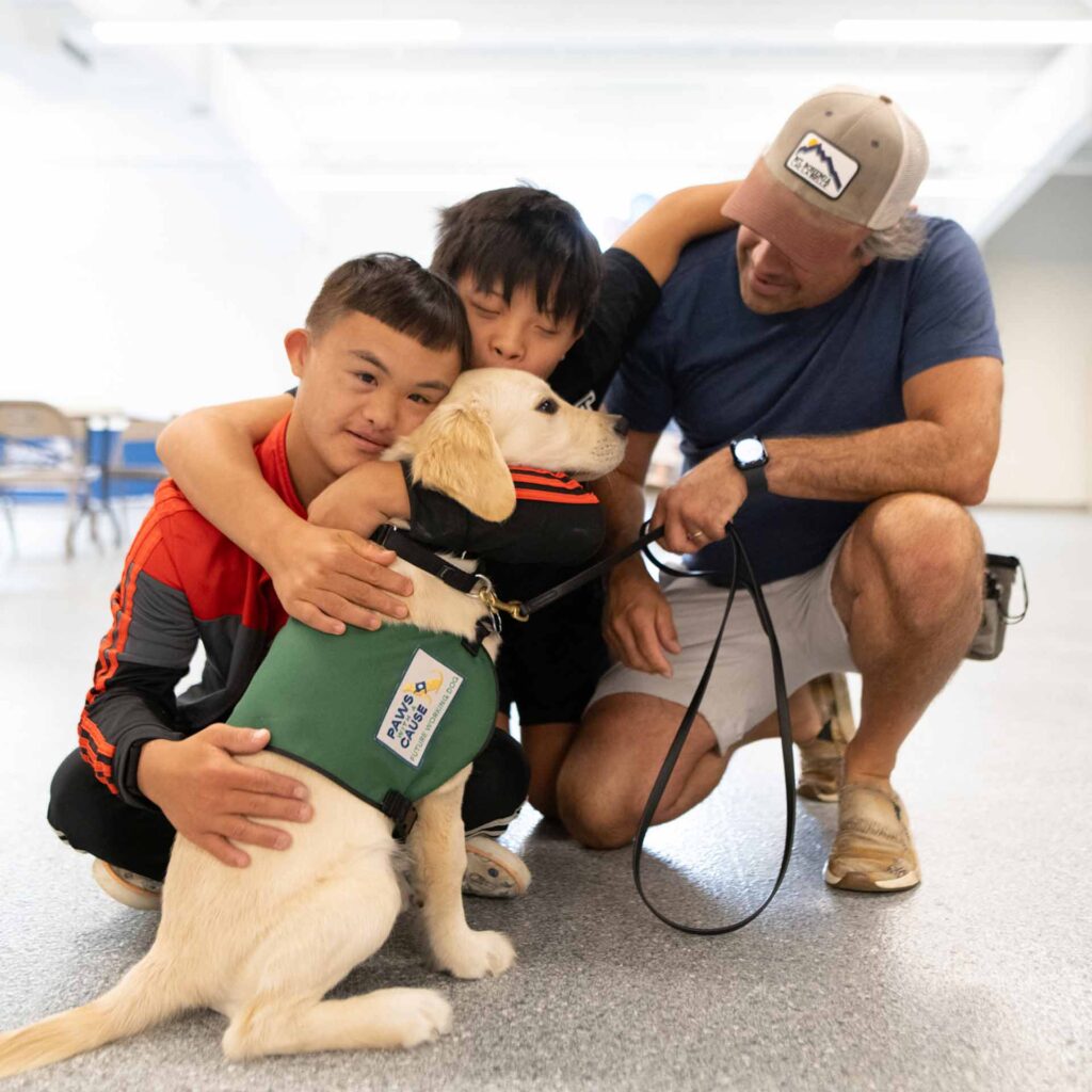 Two kids hugging a service dog with a trainer in a facility, highlighting companionship and training.