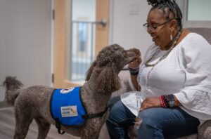 Woman smiles at a therapy dog wearing a blue assistance vest indoors.