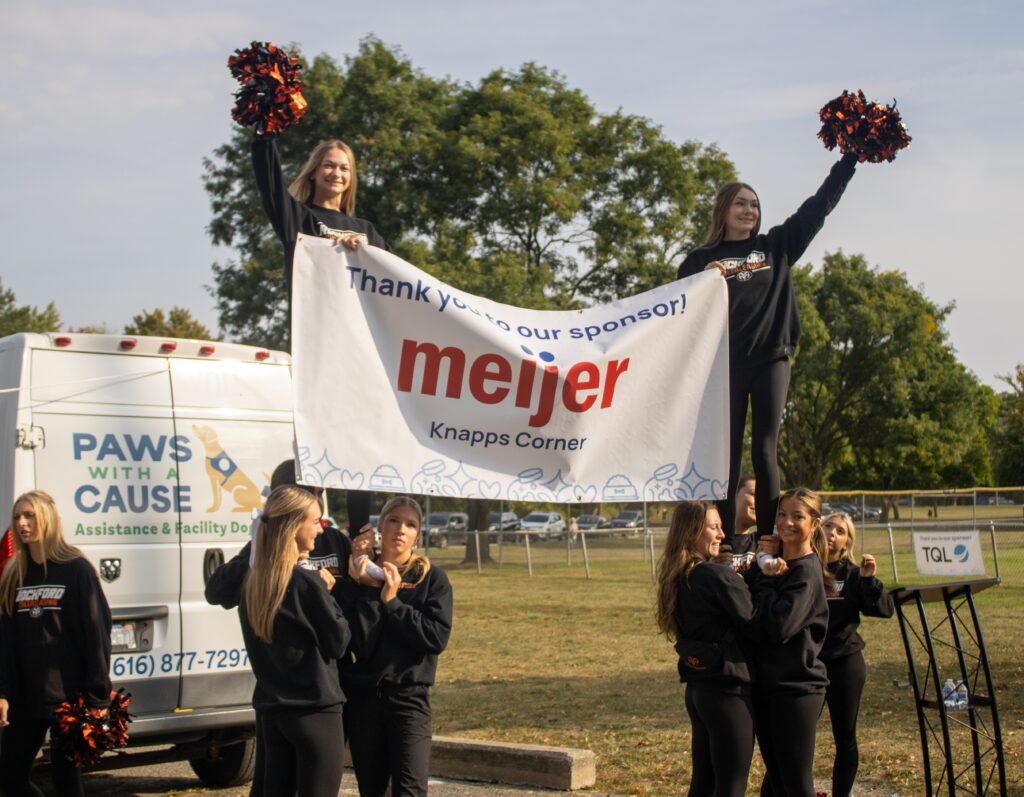 Cheerleaders holding a Meijer Knapps Corner sponsor banner at a Paws With a Cause event outdoors.