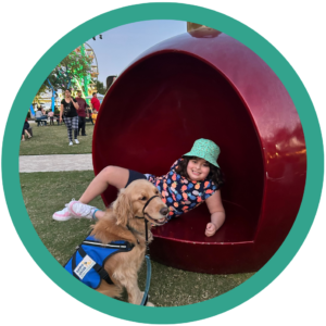 Child in a hat playing in a red sculpture, with a service dog nearby at a lively outdoor event.