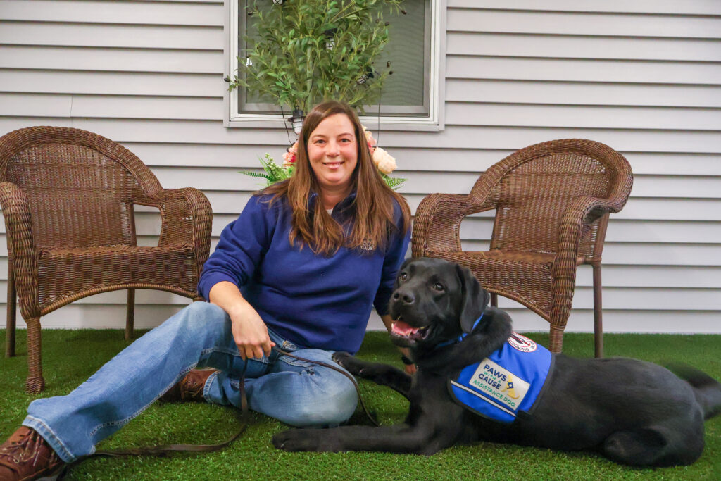 Woman sitting with black assistance dog on grass, smiling outdoors by wicker chairs.