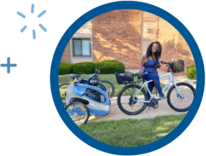 Woman smiling on a bike with a puppy trailer, parked on a sunny sidewalk in front of a brick building.