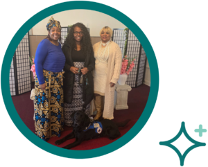 Three women smiling with a service dog at an indoor event with a floral backdrop.