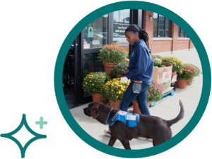 Woman and service dog entering a store, surrounded by colorful flowers in pots outside.