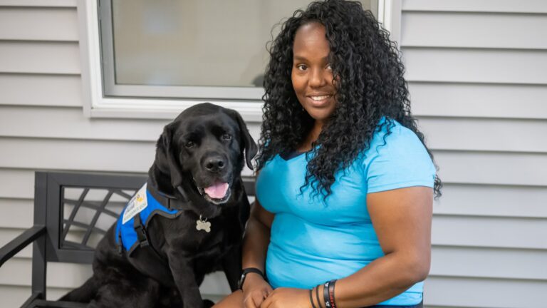Woman sitting with a black lab service dog wearing a blue vest on a bench outside.