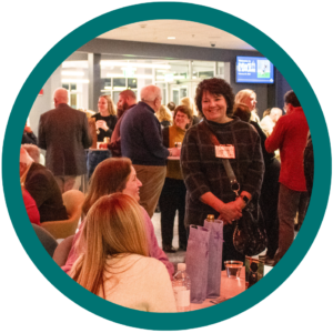 People networking at an event, with a focus on a smiling woman standing and chatting with others seated at a table.