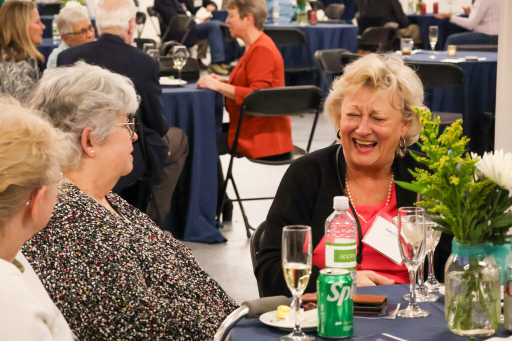 Senior women laughing and socializing at an event, enjoying drinks and conversation at a round table.
