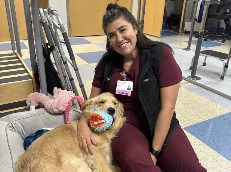 Rehab assistant sitting with a happy facility dog holding a toy, promoting therapy and companionship in a rehab center setting.