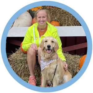 Smiling woman with golden retriever at a pumpkin patch sitting on hay, both enjoying a fall day.