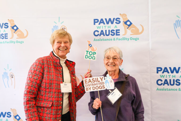 Two women smiling with dog-themed signs at a Paws With A Cause event backdrop for assistance dogs.
