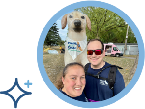 Two people smile next to a large dog statue wearing a Paws With A Cause bandana at an outdoor event.