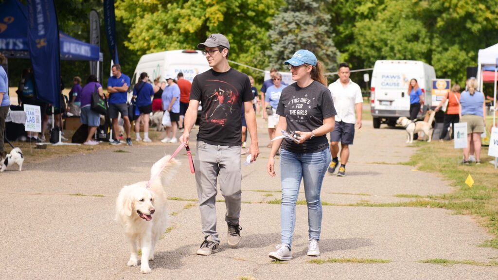 Couple walking a dog at an outdoor event with people and vendor booths in the background.