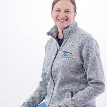 Smiling person in a gray jacket with Paws for a Cause logo, sitting on a stool against a white background.