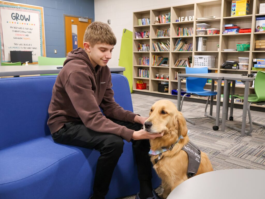 Teen sitting on a blue couch in a classroom, gently petting a golden retriever service dog.