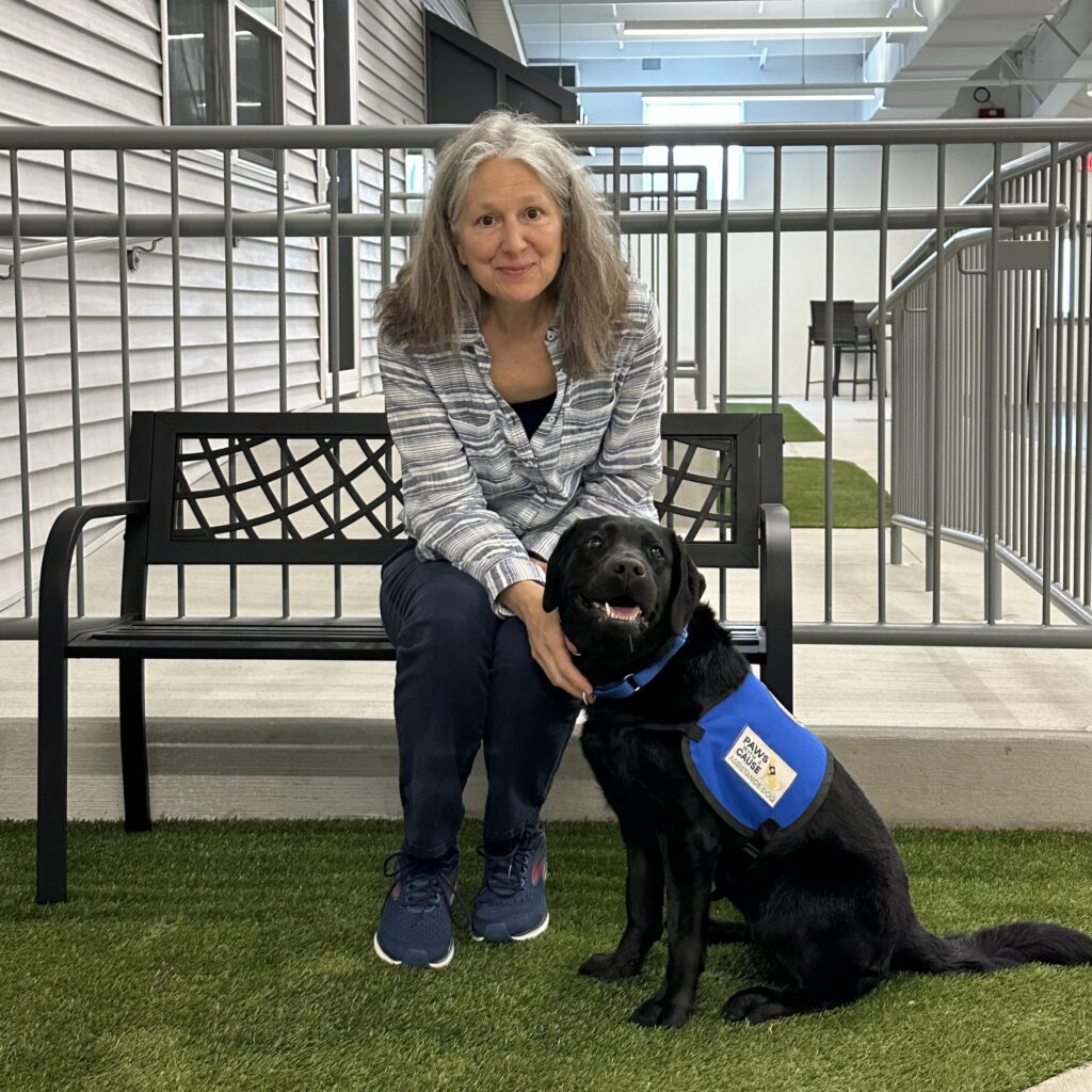 Woman sitting on bench with a black service dog wearing a blue vest inside a building with green artificial grass.
