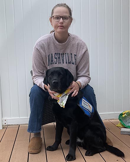 Person sitting with a black Labrador wearing a service dog vest on a wooden deck.