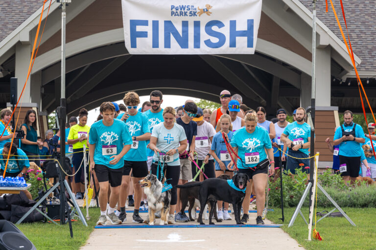 Participants and dogs at the starting line of the Paws in the Park 5K race under a finish banner.
