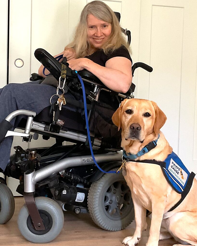 Woman in wheelchair smiling alongside a yellow service dog wearing a blue vest, indoors.