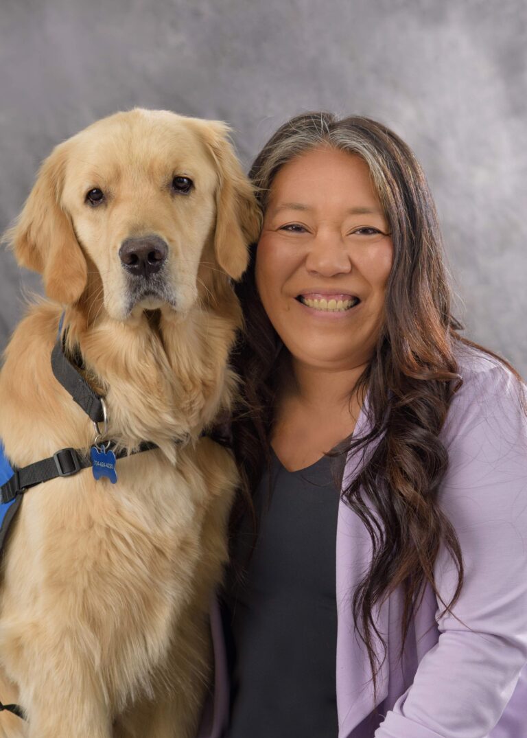 Smiling woman with long hair posing with a golden retriever wearing a blue harness against a gray background.