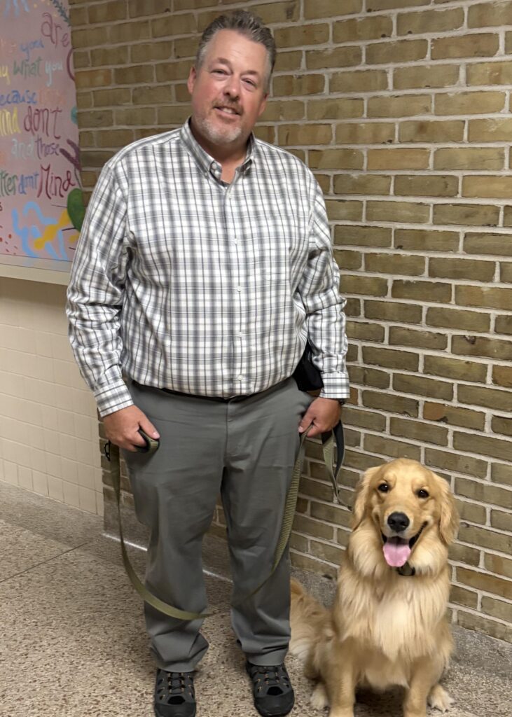 Man in plaid shirt with a golden retriever dog in hallway, brick wall background, leash in hand, smiling.
