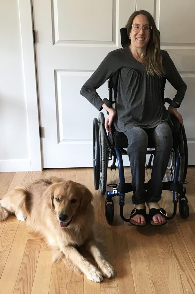 Woman in wheelchair smiling with a golden retriever on wooden floor at home.