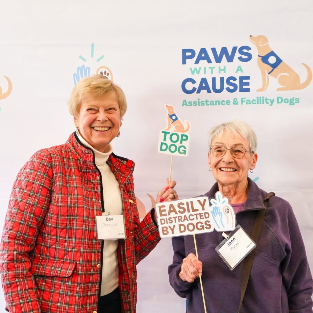 Two women smiling with Paws With a Cause backdrop, holding signs: Top Dog and Easily Distracted by Dogs.