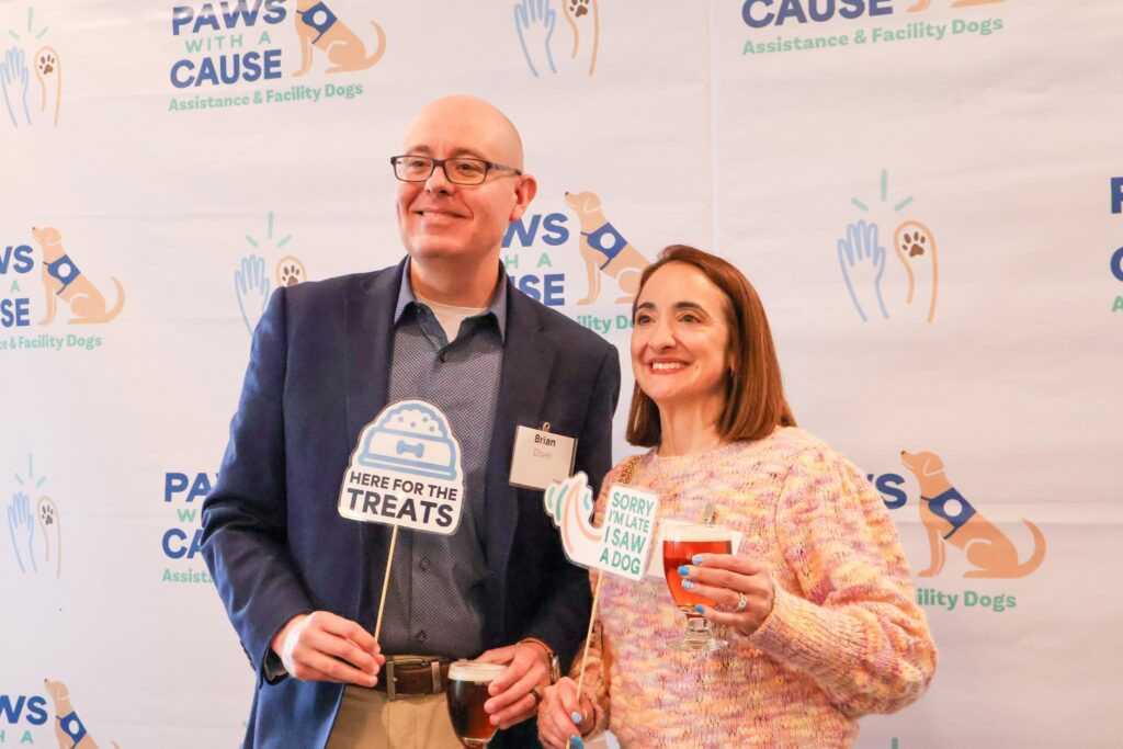 Two people smiling at a Paws With a Cause event, holding fun signs and drinks.