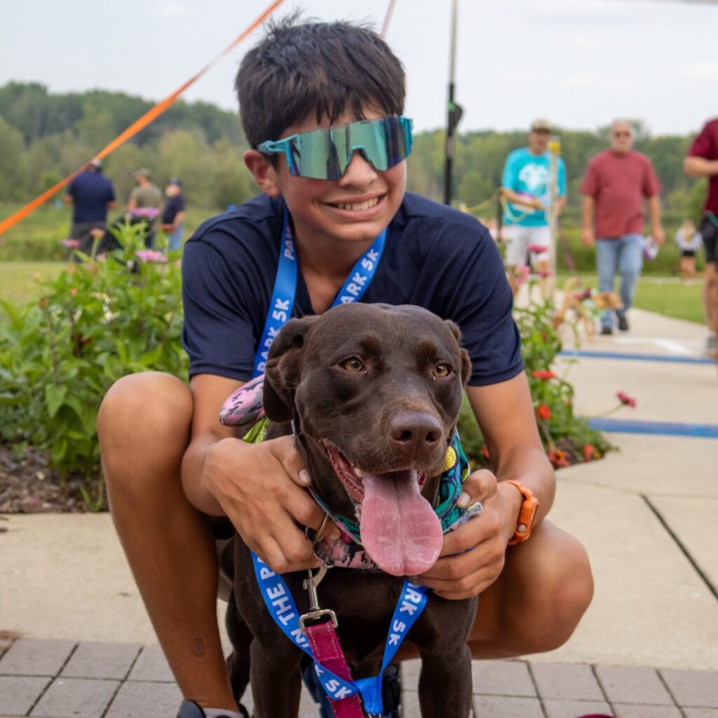 Boy in sunglasses smiling with a brown dog wearing a medal at a park event.