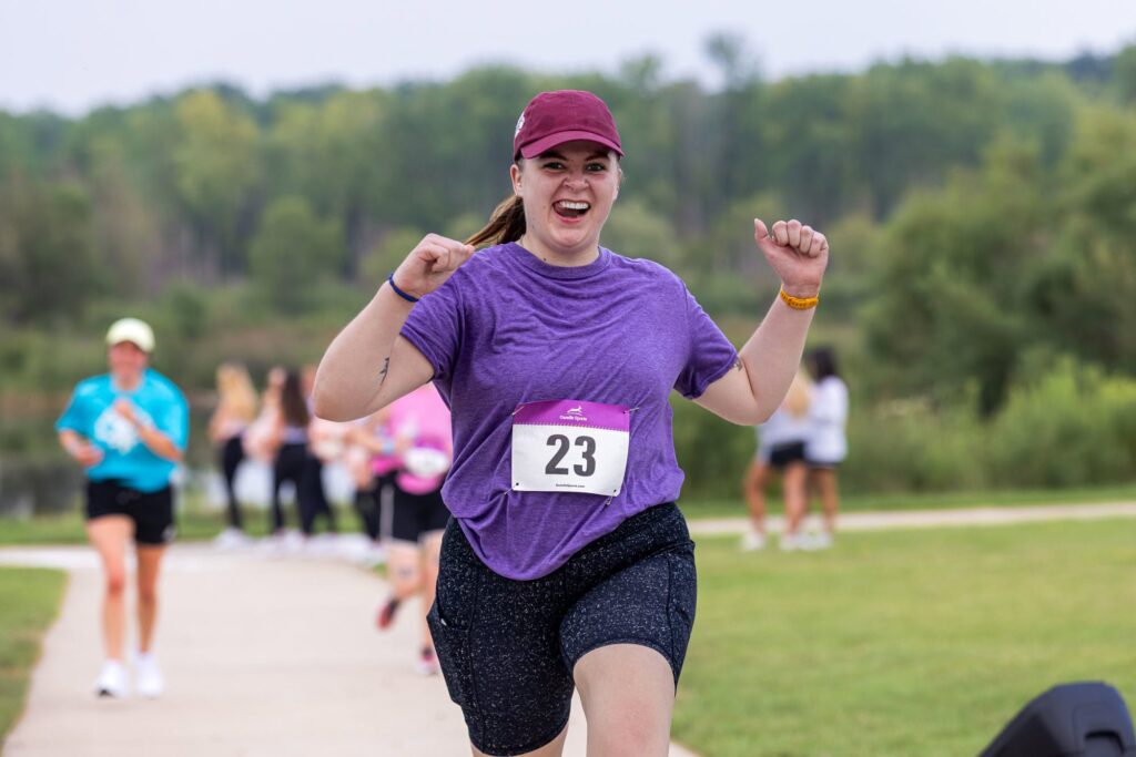 Runner finishing a race with joy in a park setting, wearing a purple shirt and bib number 23.