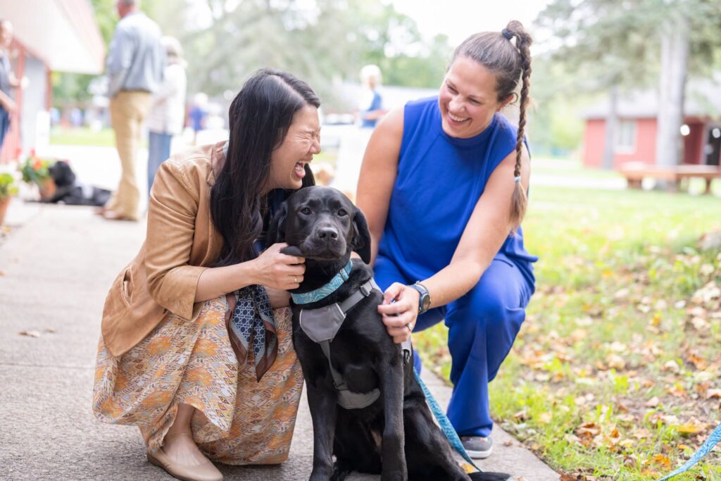 Women happily petting a black Labrador dog outdoors in a park setting.